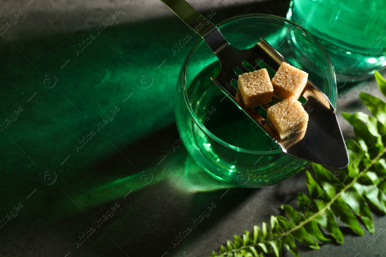 Photo of Absinthe in glass, spoon, brown sugar, and fern leaf on gray table, above view with space for text. Alcoholic drink