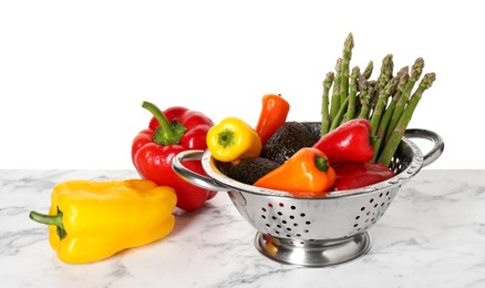 Metal colander with different vegetables and avocados on marble table against white background