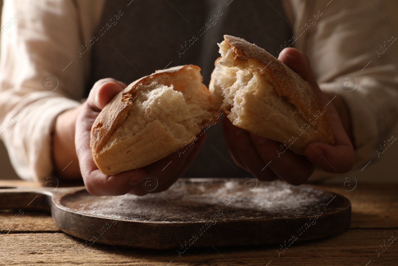 Photo of Man breaking loaf of fresh bread at wooden table, closeup