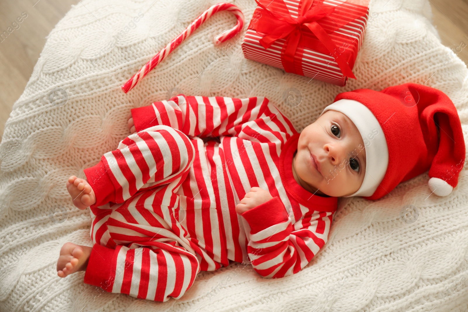 Photo of Cute little baby in Christmas hat with gift box and candy cane on knitted blanket, top view