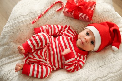 Photo of Cute little baby in Christmas hat with gift box and candy cane on knitted blanket, top view