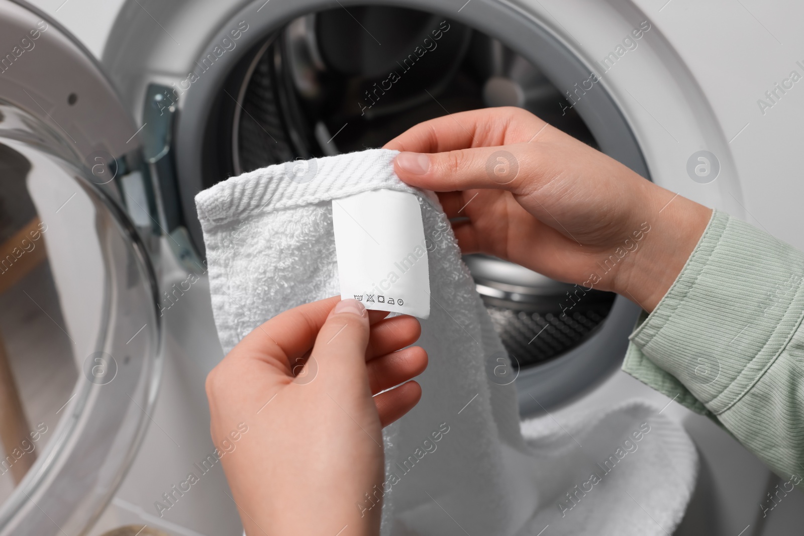 Photo of Woman reading clothing label with care symbols and material content on white towel near washing machine, closeup