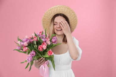 Happy young woman in straw hat holding bouquet of beautiful tulips on pink background