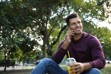 Photo of Handsome man with refreshing drink talking on smartphone in park