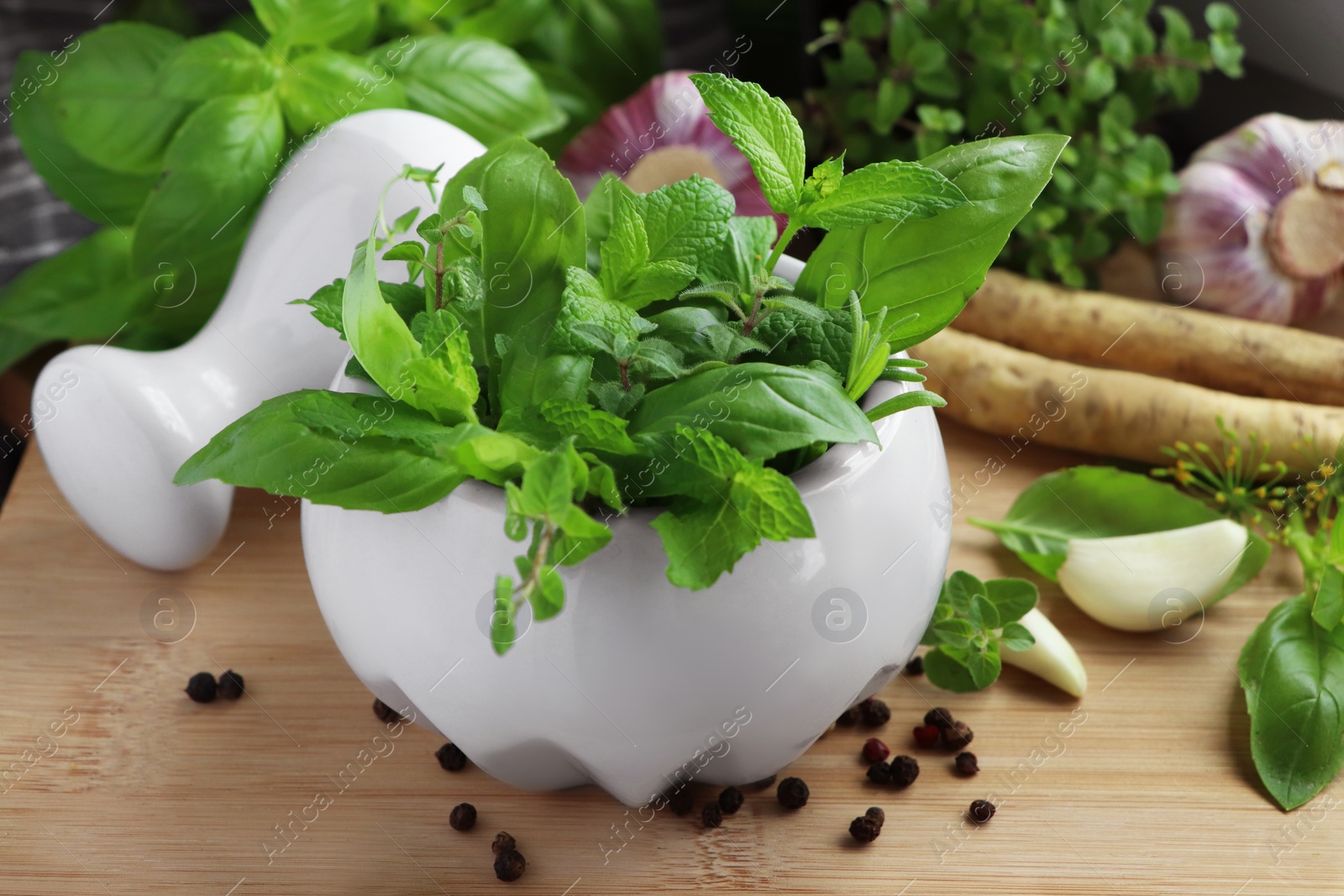 Photo of Mortar with different fresh herbs near garlic, horseradish roots and black peppercorns on wooden table, closeup