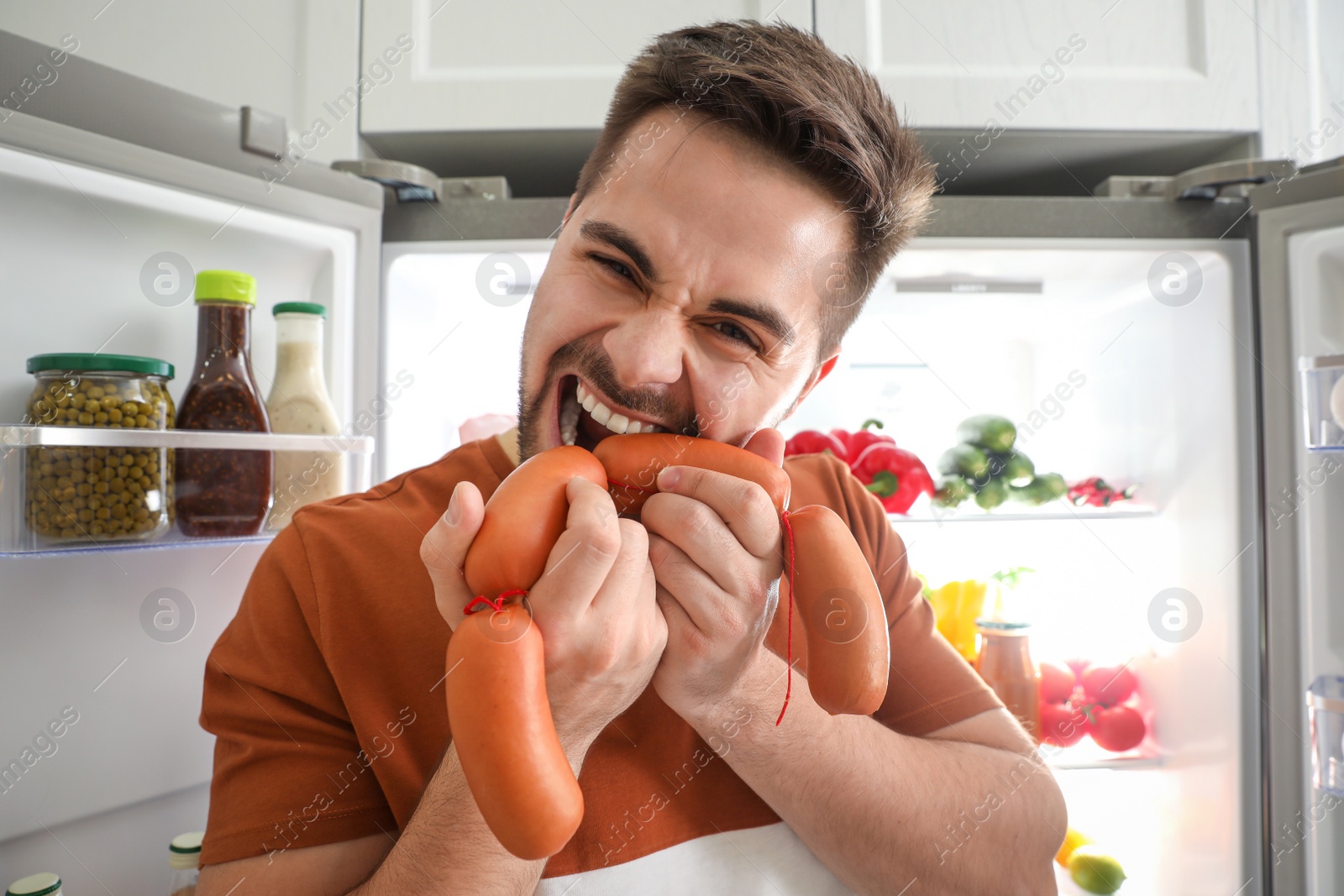 Photo of Young man eating sausages near open refrigerator indoors