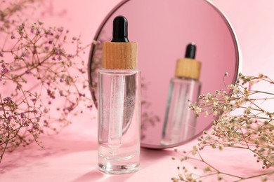 Photo of Bottle of face serum and beautiful flowers near mirror on pink background, closeup
