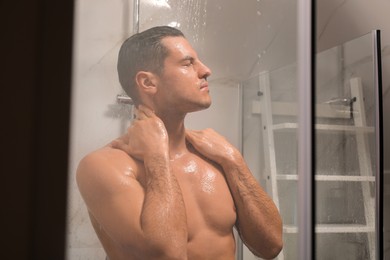 Handsome man taking shower at home, view through glass