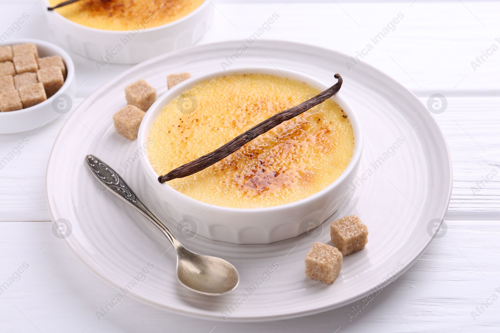 Photo of Delicious creme brulee in bowl, vanilla pod, sugar cubes and spoon on white wooden table