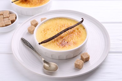 Delicious creme brulee in bowl, vanilla pod, sugar cubes and spoon on white wooden table