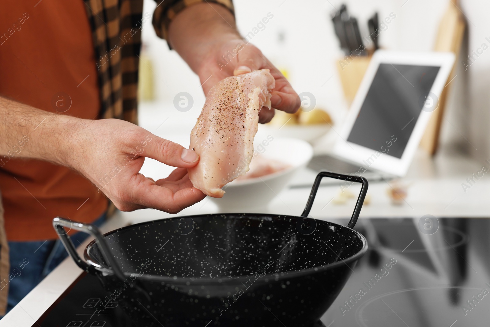 Photo of Man putting chicken fillet into frying pan while watching online cooking course via laptop in kitchen, closeup