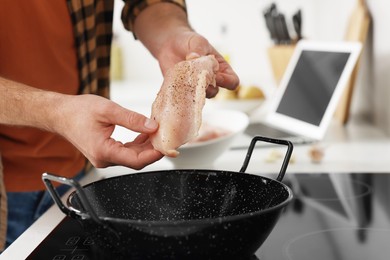 Man putting chicken fillet into frying pan while watching online cooking course via laptop in kitchen, closeup