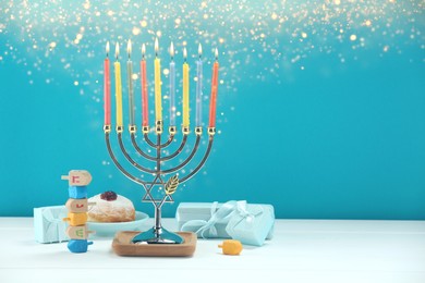 Hanukkah celebration. Menorah with burning candles, dreidels, gift boxes and donut on white wooden table against light blue background, space for text