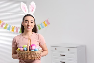 Happy woman in bunny ears headband holding wicker basket of painted Easter eggs indoors. Space for text