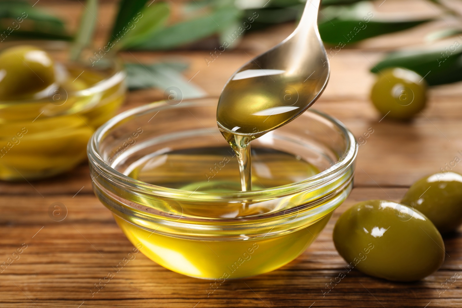 Photo of Spoon with cooking oil over bowl and olives on wooden table, closeup