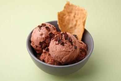 Tasty chocolate ice cream and piece of waffle cone in bowl on light green background, closeup