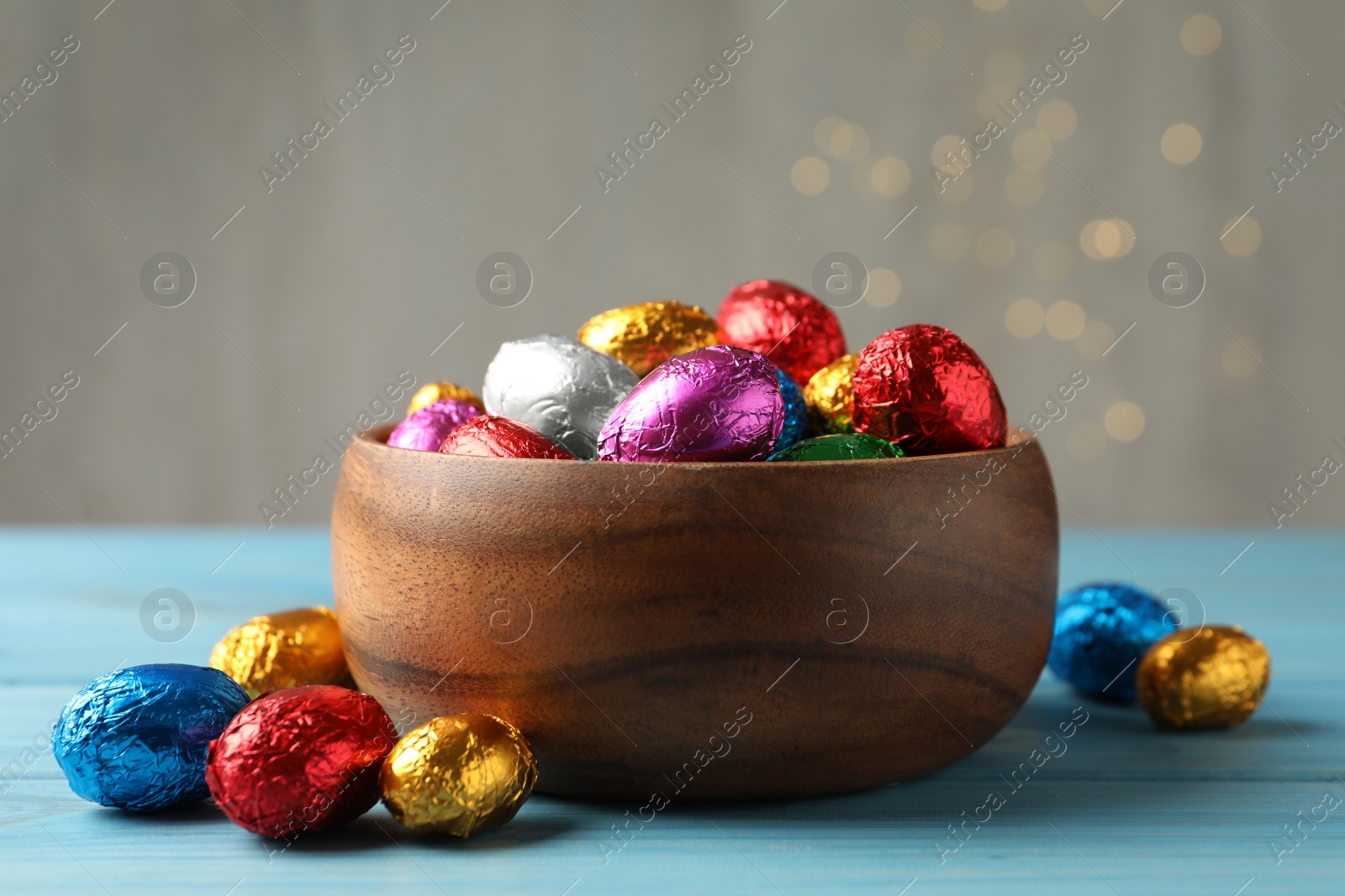 Photo of Bowl with chocolate eggs wrapped in colorful foil on light blue wooden table