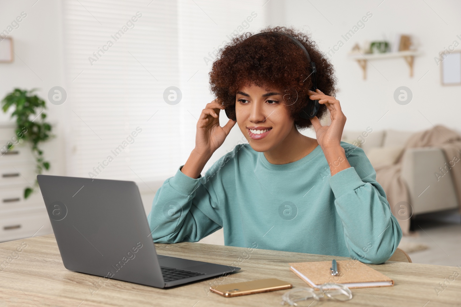 Photo of Young woman in headphones using laptop at wooden desk in room