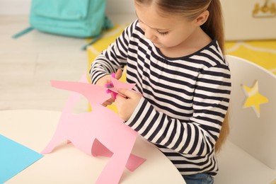 Photo of Cute little girl cutting pink paper at desk in room, closeup. Home workplace