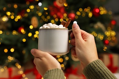 Woman holding cup of delicious cocoa with marshmallows near Christmas tree at home, closeup