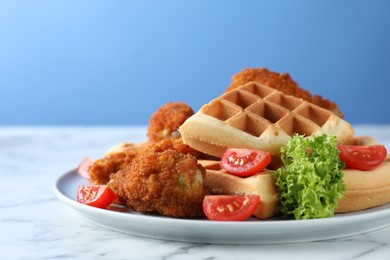 Tasty Belgian waffles served with fried chicken, tomatoes and lettuce on white marble table against light blue background, closeup