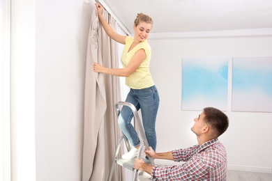 Photo of Young couple hanging window curtain indoors. Interior decor element