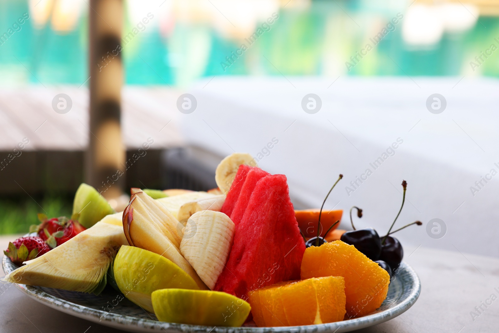 Photo of Plate with fresh fruits on table near sun lounger. Luxury resort with outdoor swimming pool