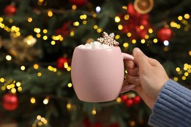 Woman holding cup of delicious cocoa with marshmallows and gingerbread cookie near Christmas tree at home, closeup