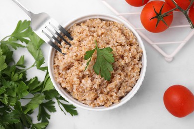 Tasty wheat porridge with parsley and tomatoes in bowl on white table, flat lay