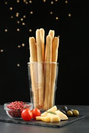 Photo of Fresh delicious grissini sticks in glass, red peppercorns, tomatoes, olives and cheese on dark table against black background