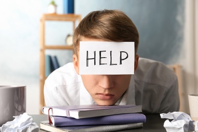 Photo of Young man with note HELP on forehead at workplace