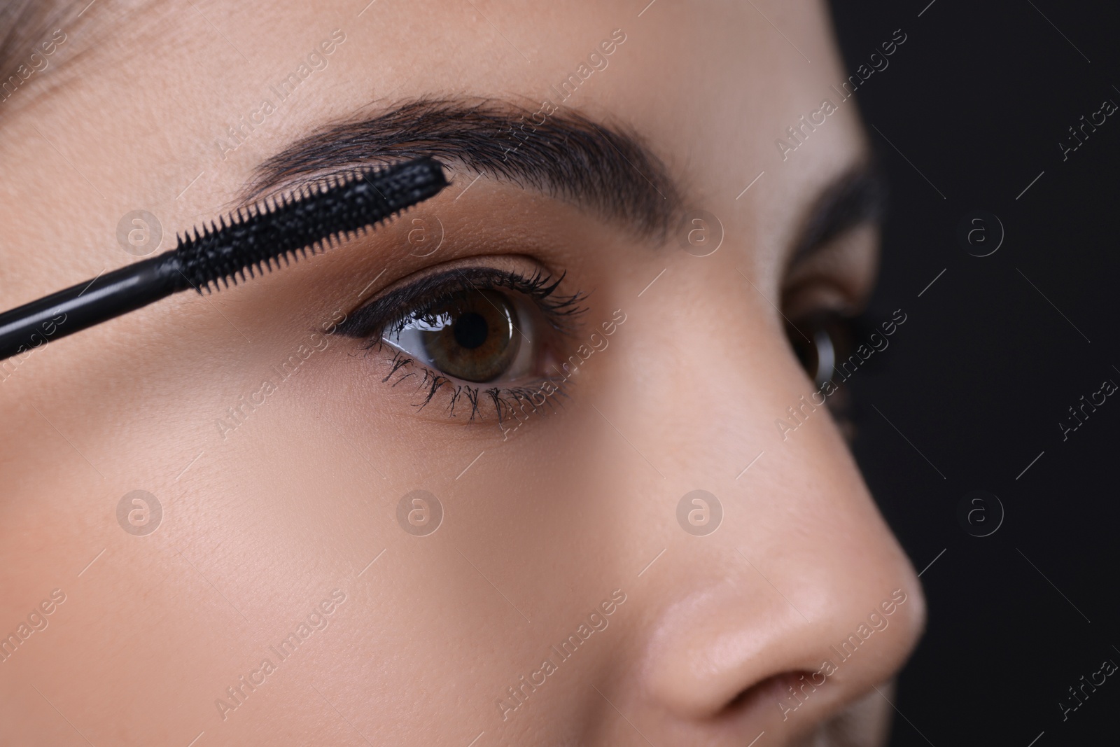 Photo of Beautiful young woman applying mascara on black background, closeup