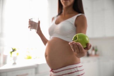 Photo of Young pregnant woman holding glass of water and apple in kitchen, focus on hand with fruit. Taking care of baby health