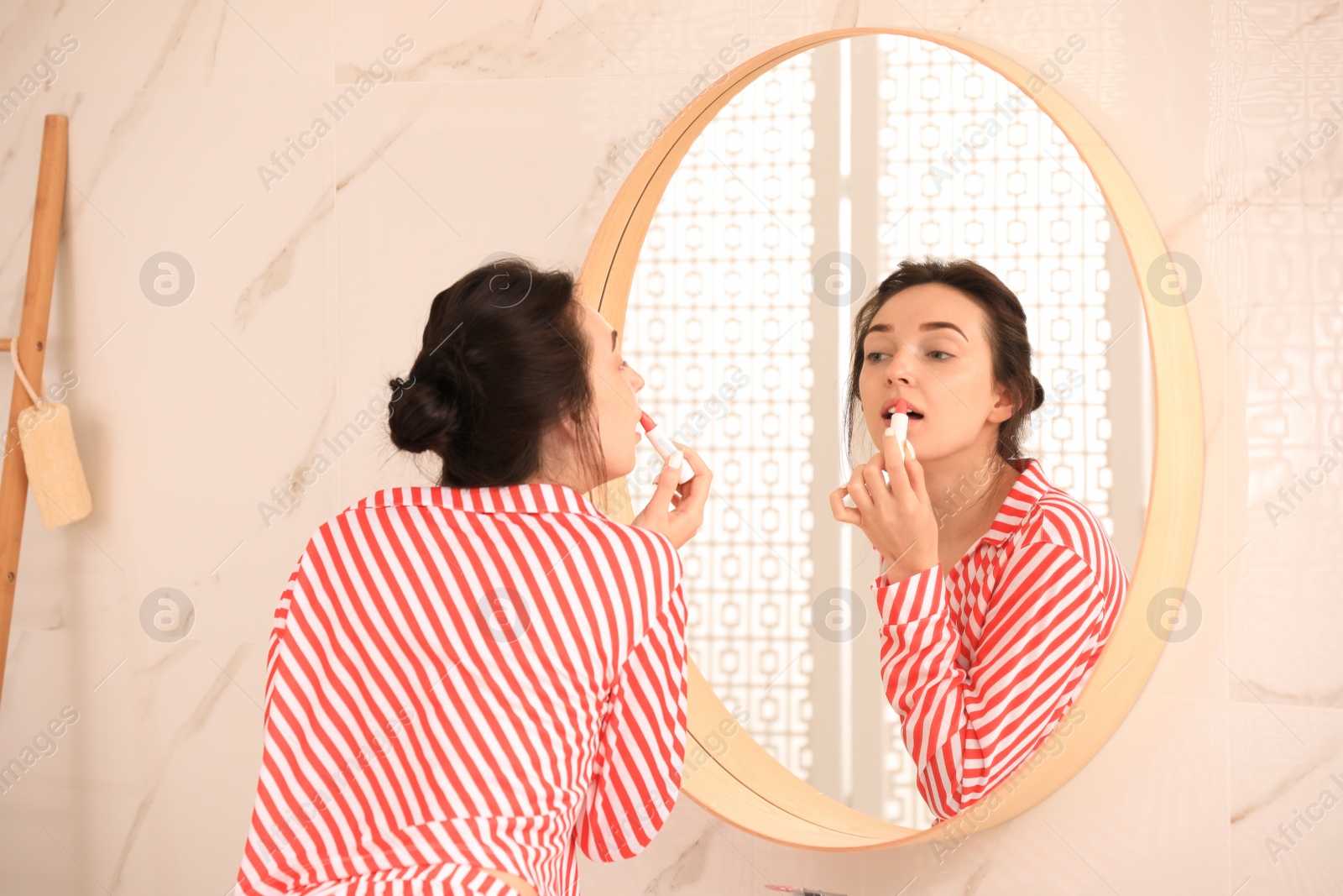 Photo of Young woman applying balm on lips near mirror in bathroom