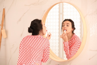 Young woman applying balm on lips near mirror in bathroom