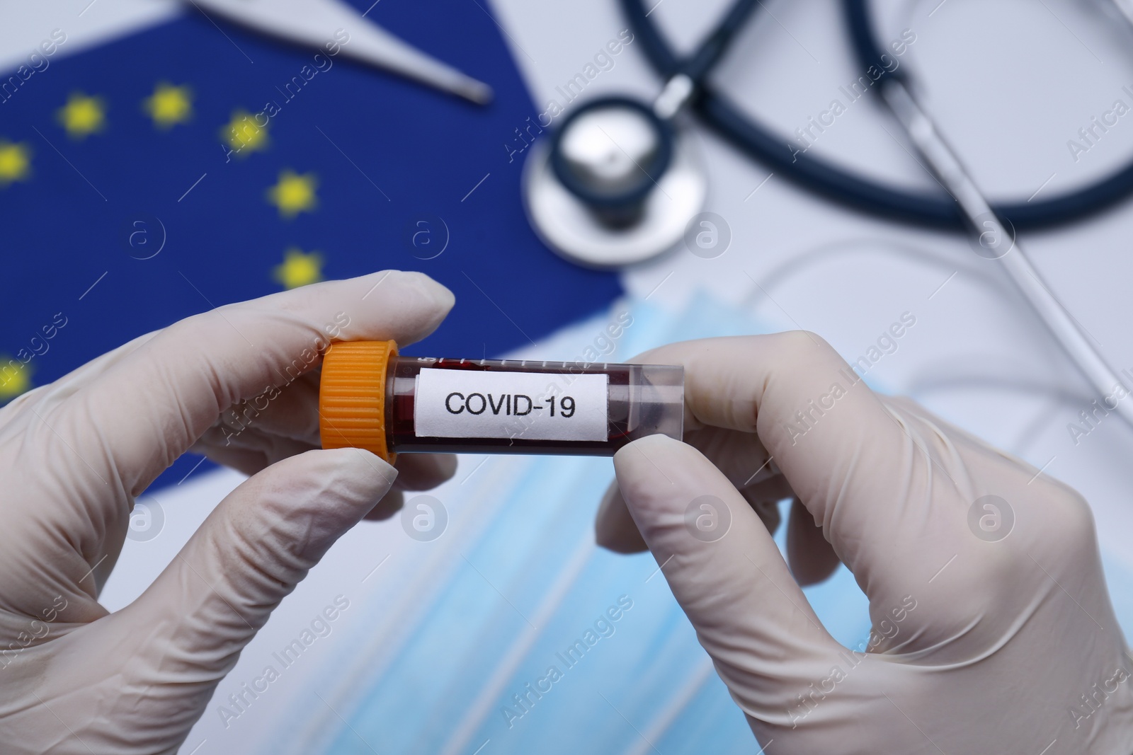 Photo of Doctor holding sample tube with label COVID-19 above medical items and European Union flag, closeup