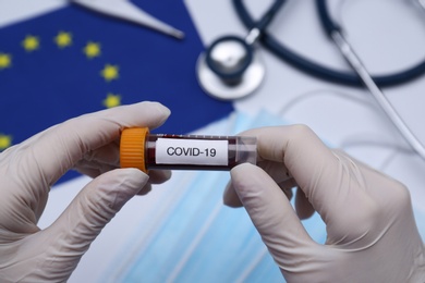 Photo of Doctor holding sample tube with label COVID-19 above medical items and European Union flag, closeup