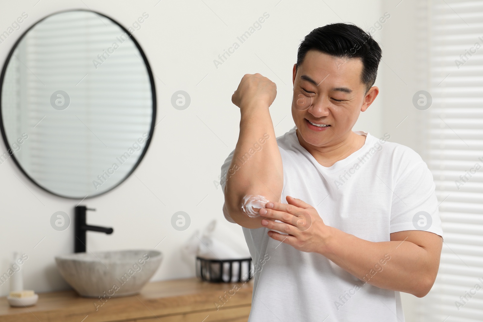 Photo of Handsome man applying body cream onto his elbow in bathroom. Space for text