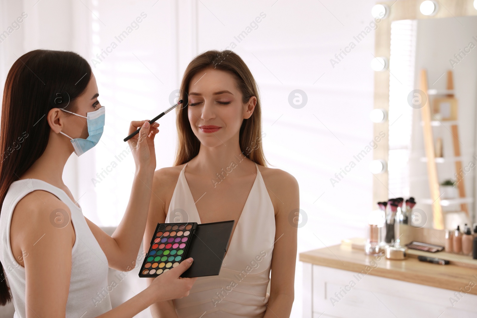 Photo of Makeup artist in protective mask working with woman indoors. Preventive measures during COVID-19 pandemic