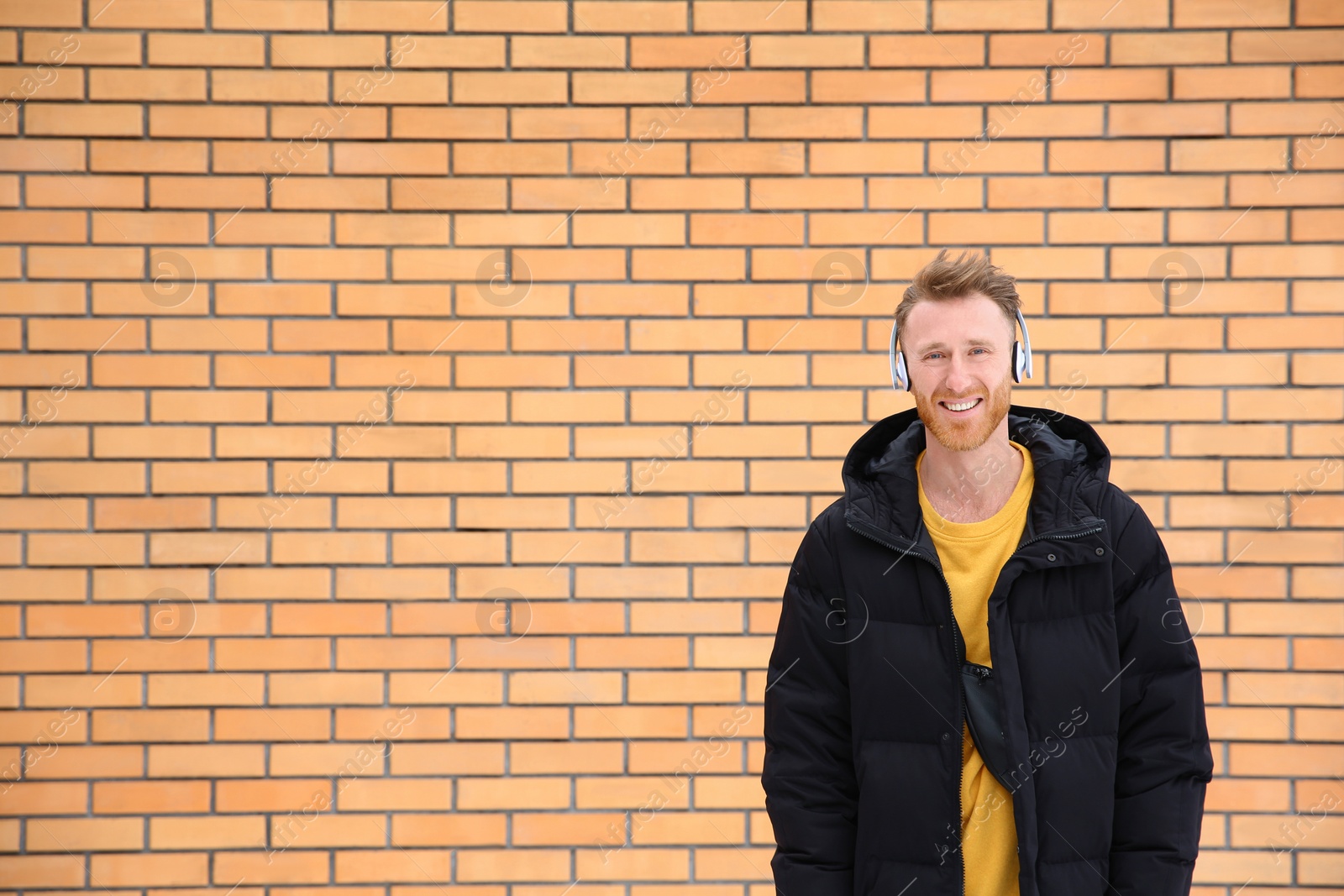 Photo of Young man listening to music with headphones against brick wall. Space for text