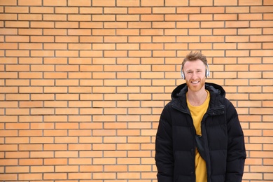 Photo of Young man listening to music with headphones against brick wall. Space for text