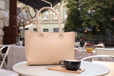 Photo of Stylish bag and cup of coffee on white table in outdoor cafe