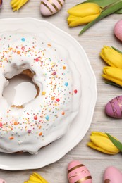 Easter cake with sprinkles, painted eggs and tulips on white wooden table, flat lay