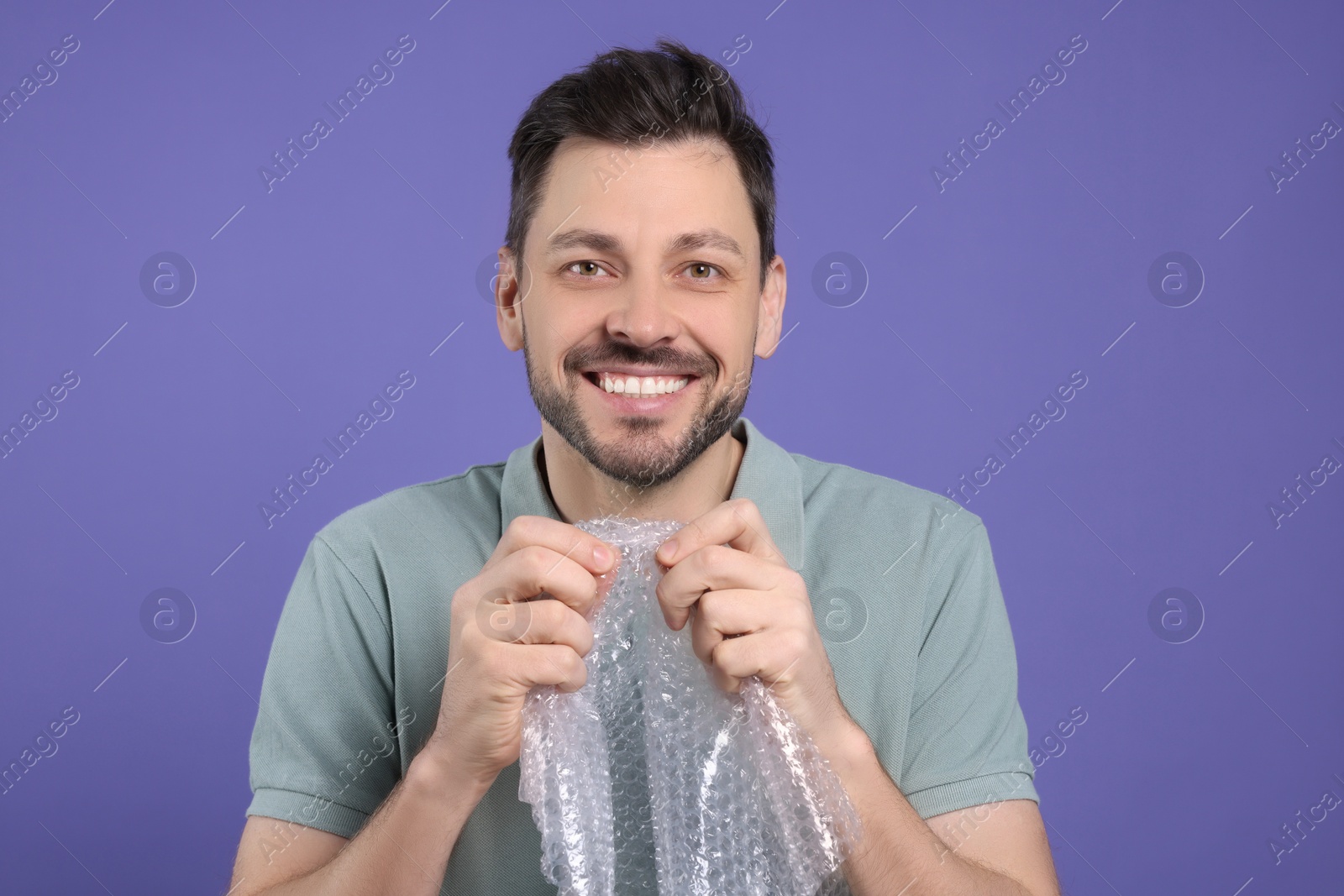 Photo of Happy man popping bubble wrap on purple background. Stress relief
