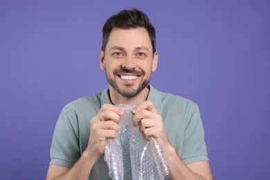 Happy man popping bubble wrap on purple background. Stress relief