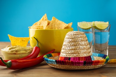 Photo of Mexican sombrero hat, tequila, chili peppers, nachos chips and guacamole on wooden table against light blue background