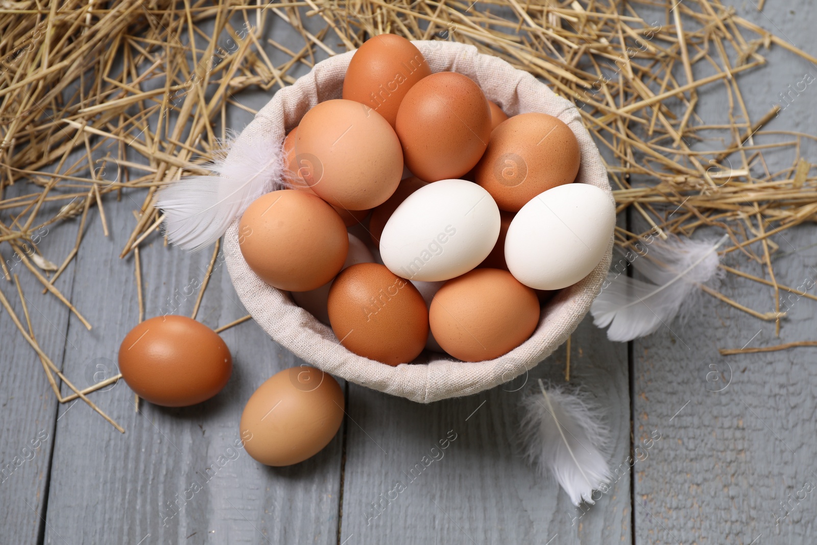 Photo of Fresh chicken eggs in bowl, feathers and dried straw on grey wooden table, above view