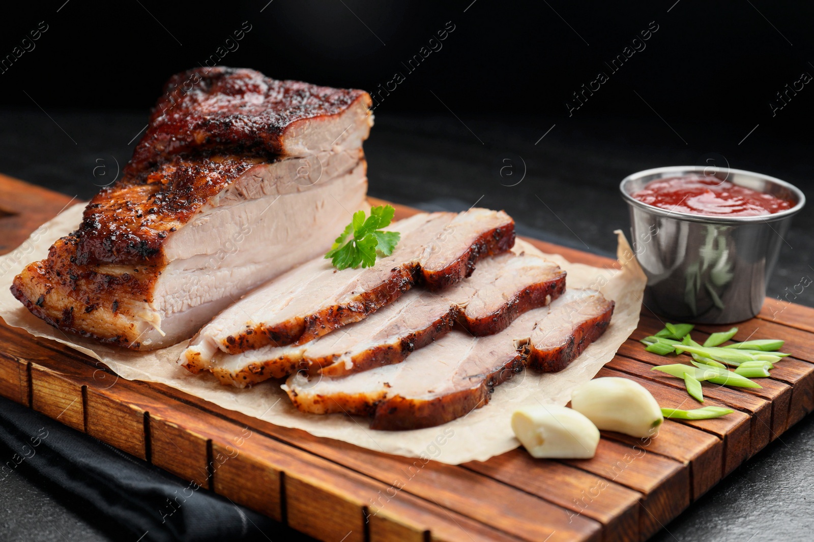 Photo of Pieces of baked pork belly served with sauce and parsley on black textured table, closeup