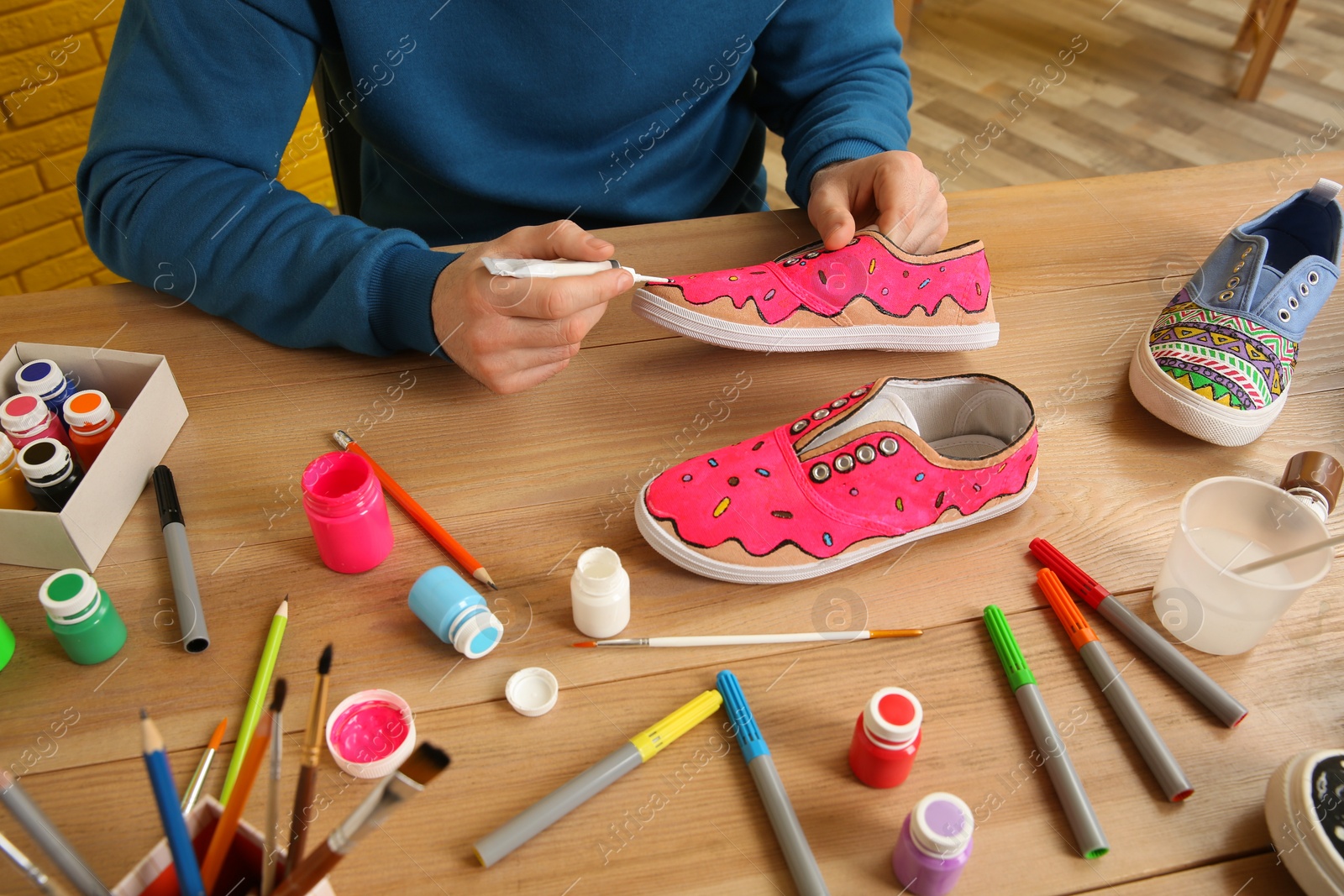 Photo of Man painting on sneaker at wooden table indoors, closeup. Customized shoes