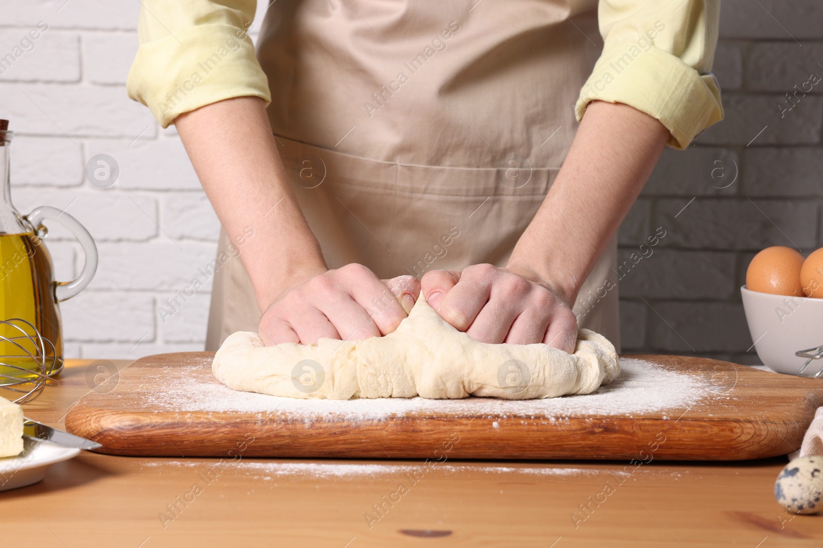 Photo of Woman kneading dough at wooden table near white brick wall, closeup